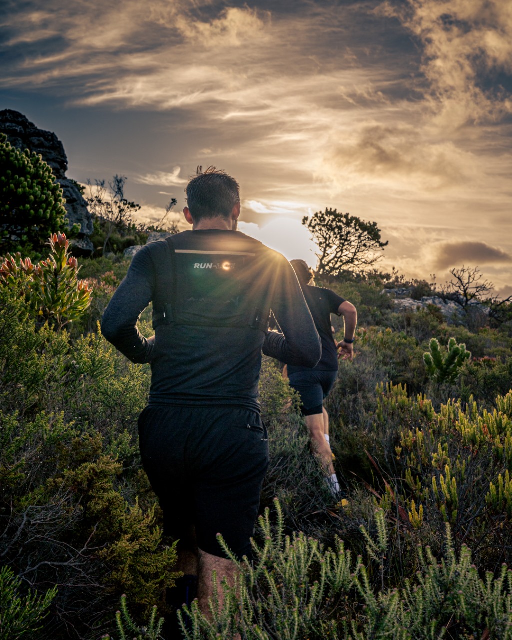Two trail runners navigating fynbos with dramatic sun rays and mountain backdrop