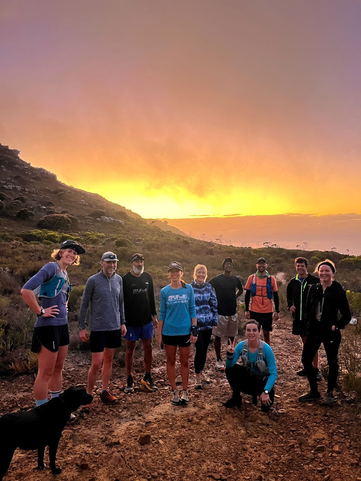 Large group of TrailDawgs posing together on mountain trail during spectacular sunset