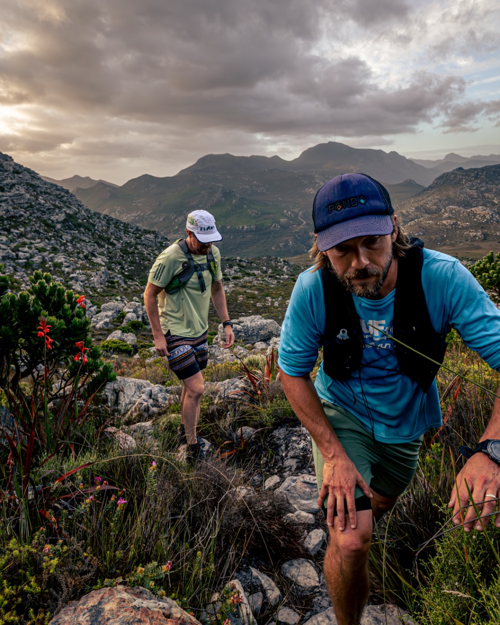 Two trail runners on rocky mountain terrain during golden hour with dramatic mountain silhouettes