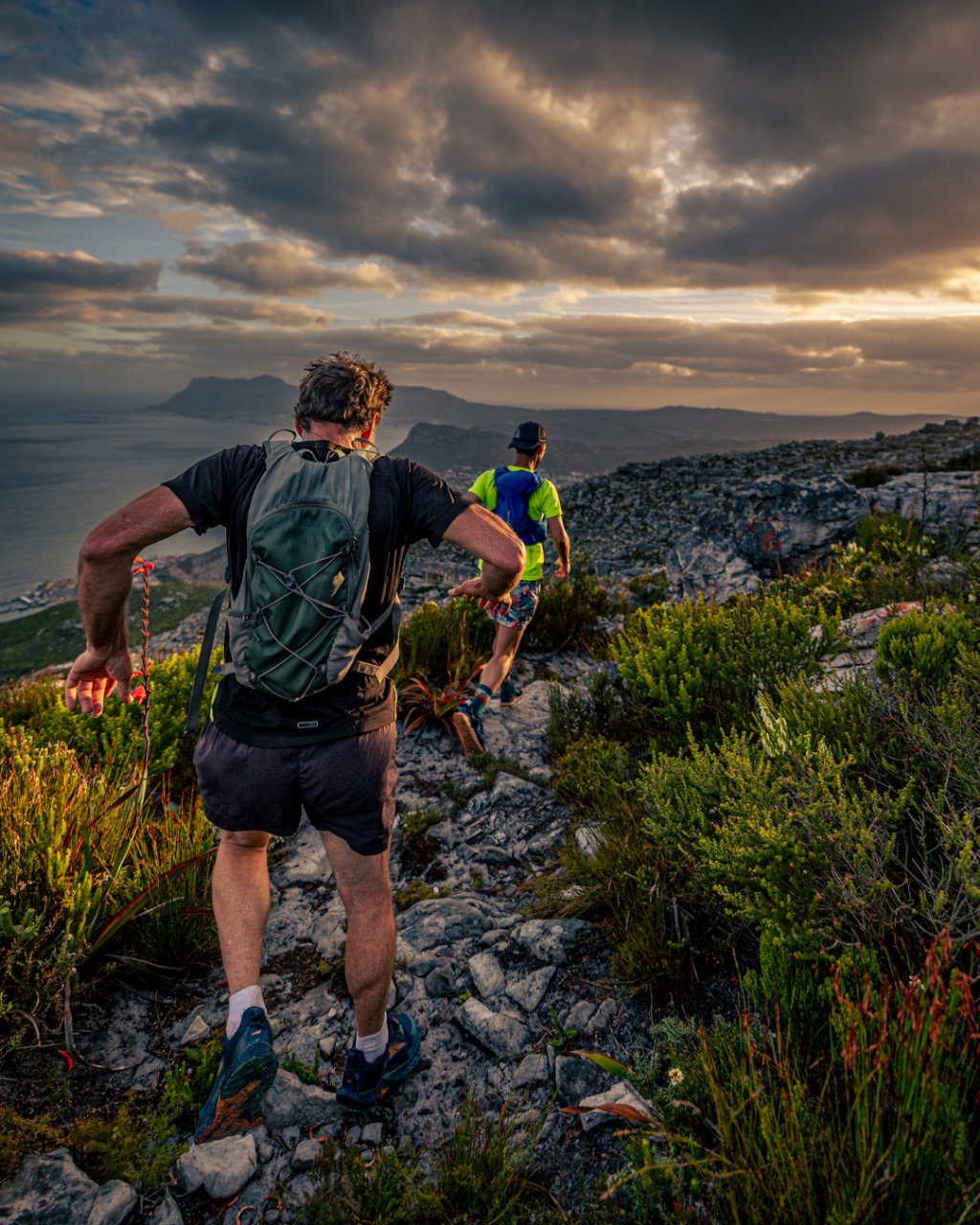 Two trail runners on rocky mountain terrain with dramatic cloudy sky and ocean views