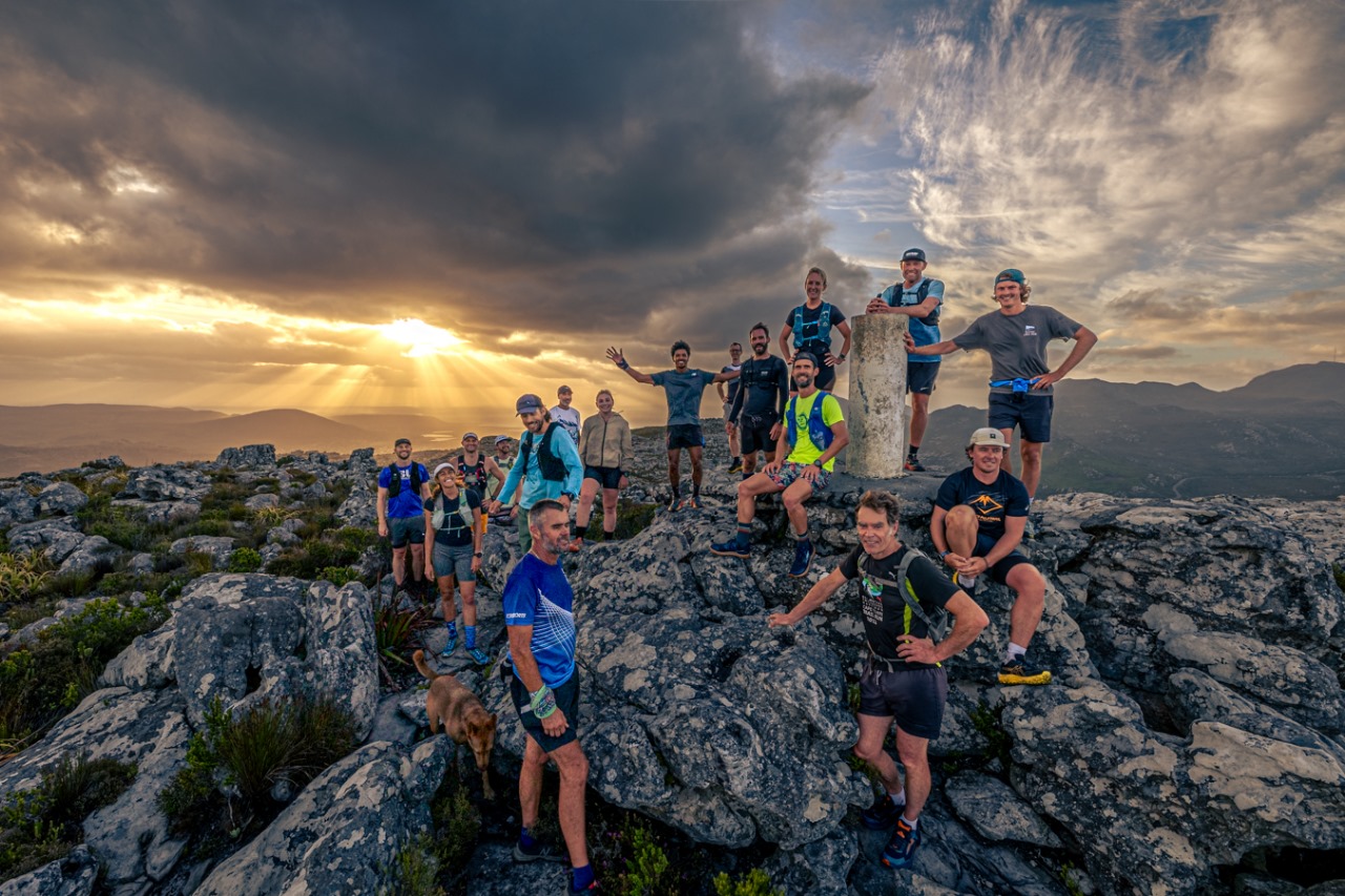 TrailDawgs group gathered around trig beacon during golden hour sunset on mountain peak